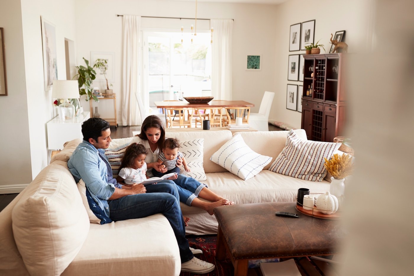 Young family sitting on sofa reading a book together in the living room, seen from doorway