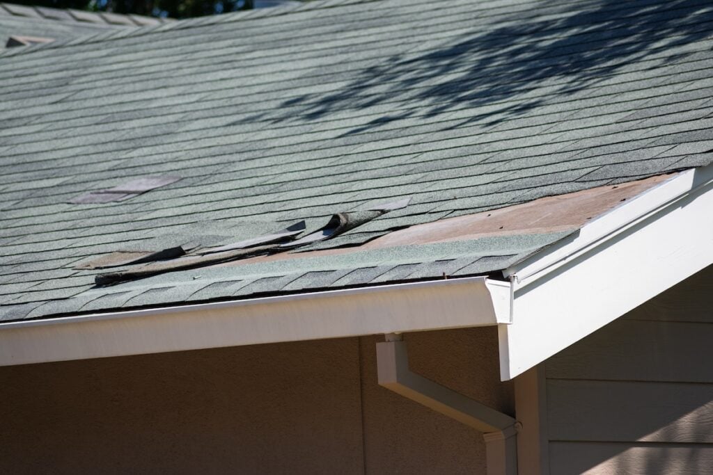 Asphalt shingle roofing on a residential home with visible damage, curled edges, and exposed sheathing.