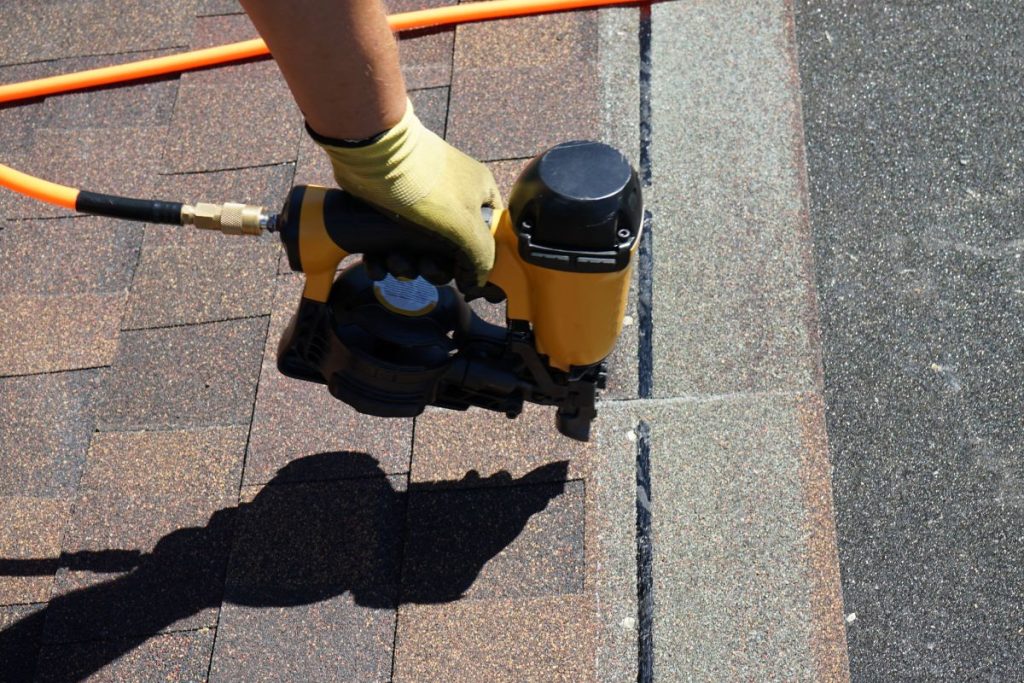 Roofer builder worker with nailgun installing Asphalt Shingles or Bitumen Tiles on a roof of a new house